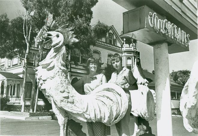 Two people pose on a rooster statue near a sign and a Victorian-style building.