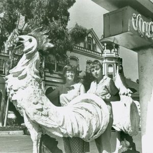 Two people pose on a rooster statue near a sign and a Victorian-style building.