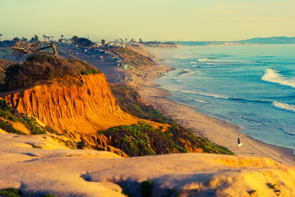 Coastal landscape with cliffs, sandy beach, and ocean waves at sunset.