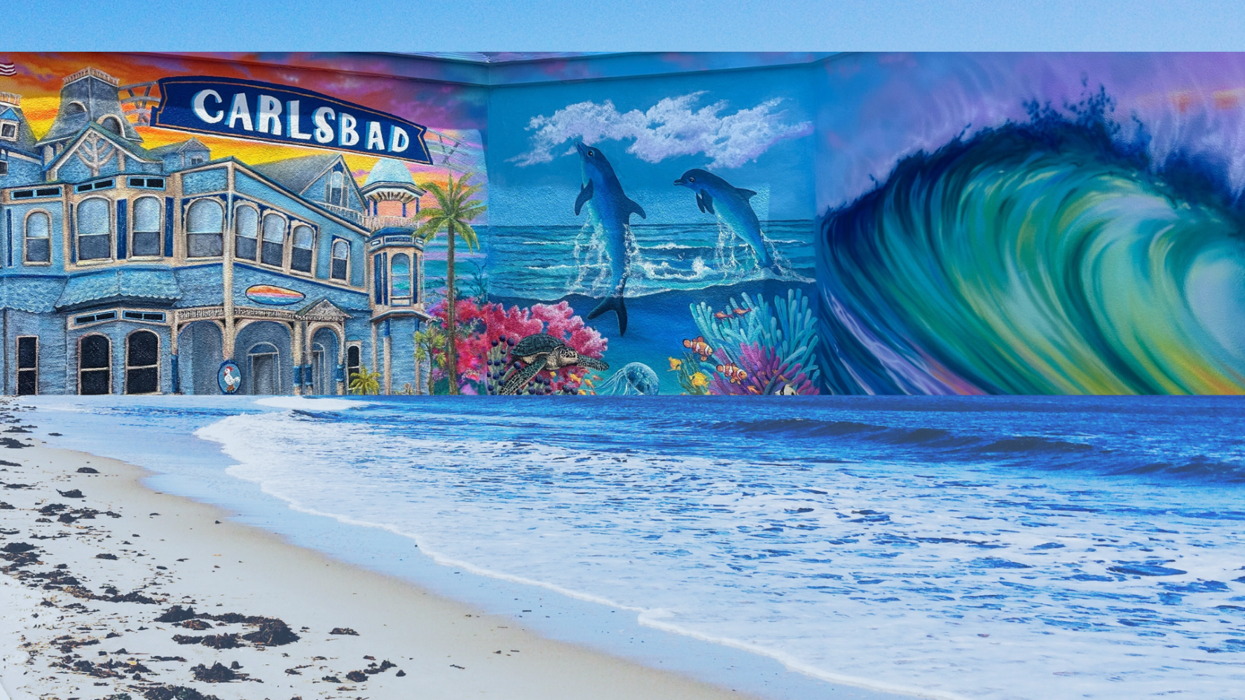 Colorful mural of Carlsbad with ocean, dolphins, and wave scene on beach wall.