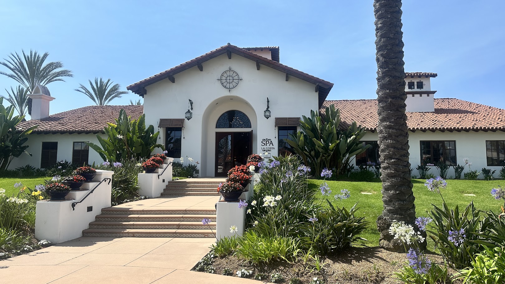 Elegant building with red tile roof and palm trees, surrounded by landscaped gardens.