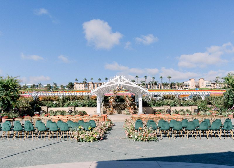 Outdoor wedding setup with chairs facing white archway under blue sky.