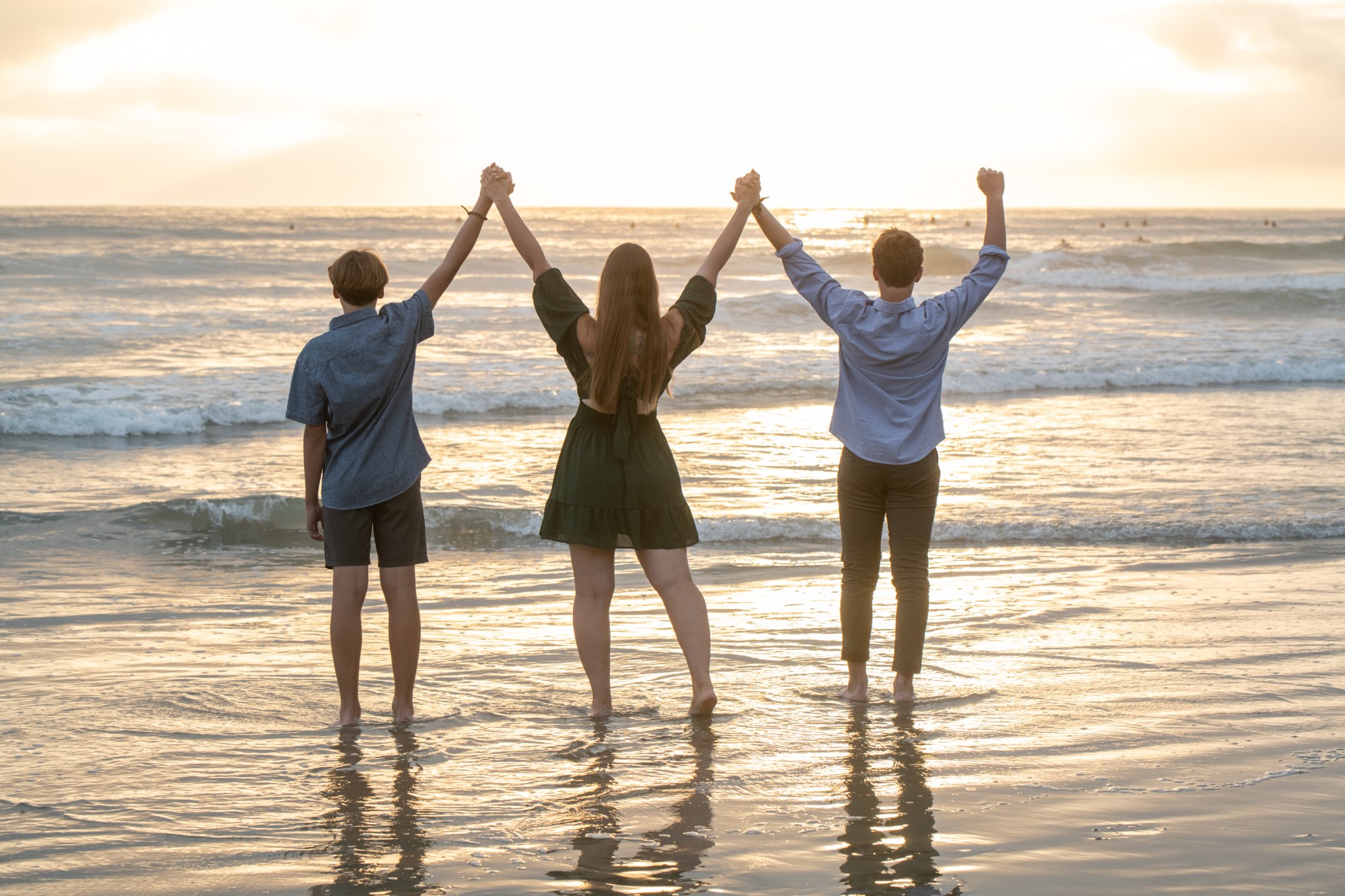 Three people holding hands, standing in ocean waves facing sunset.