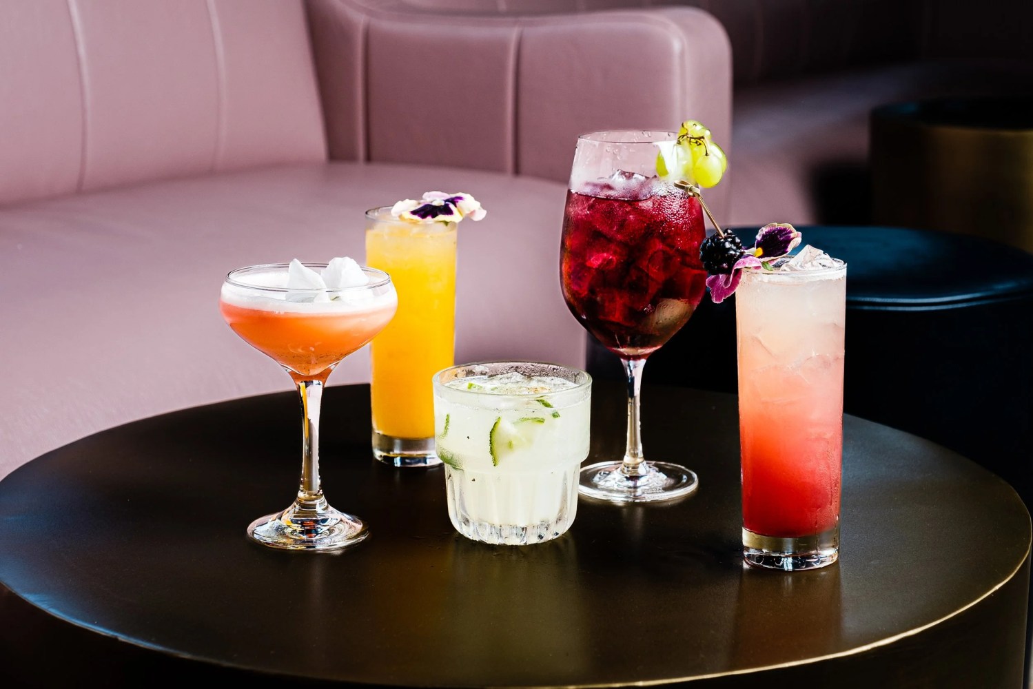 Five vibrant cocktails on a dark table with a pink sofa background.