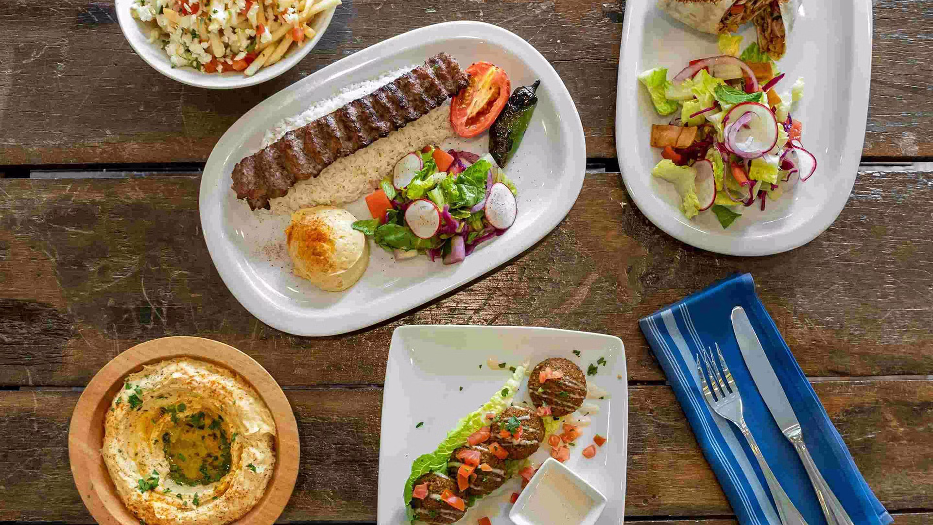 Plates of Mediterranean food including kebabs, salad, hummus, and rice on a wooden table.
