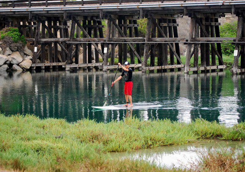 Person paddleboarding on calm water near a wooden bridge.