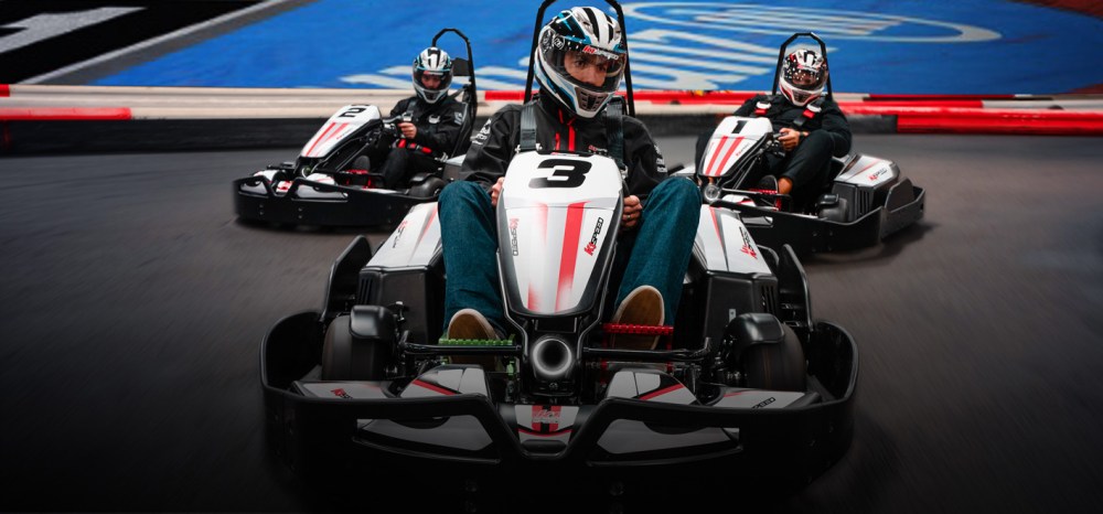 Three people racing go-karts on an indoor track, wearing helmets and racing gear.