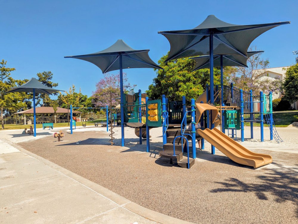 Sunny playground with slides, swings, and shade canopies in a park setting.