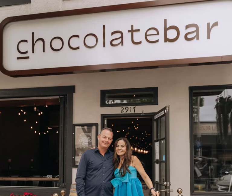 Two people smiling in front of 'chocolatebar' store sign.