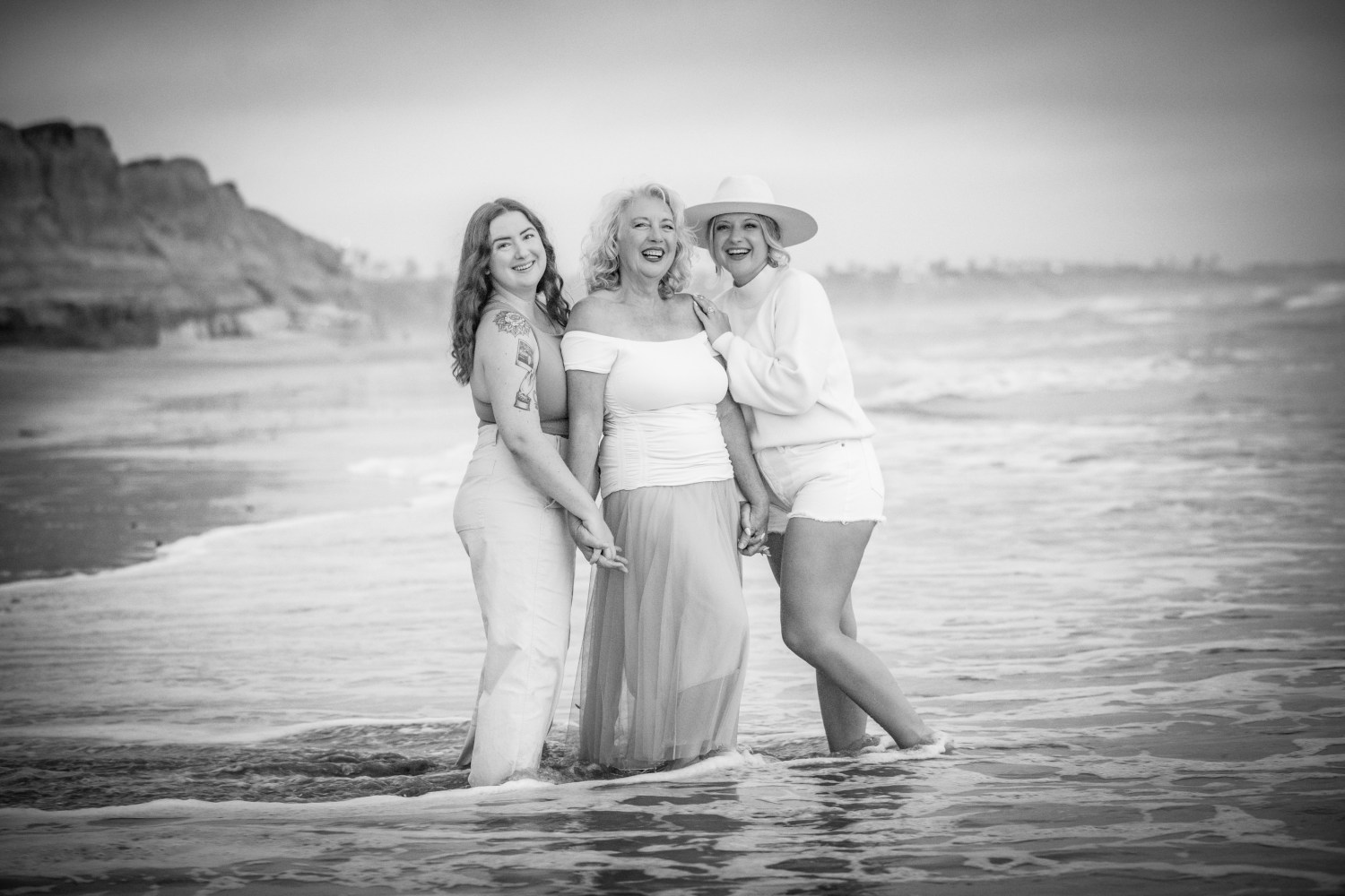 Three women smiling and standing in shallow ocean water on a beach.