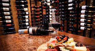Wine cellar with bottles on shelves, a wine glass, and a meat and cheese platter on a wooden table.
