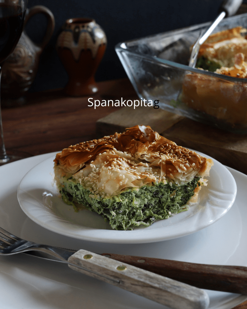 Plate of spanakopita with sesame seeds, glass baking dish, fork, knife, and pottery cup.