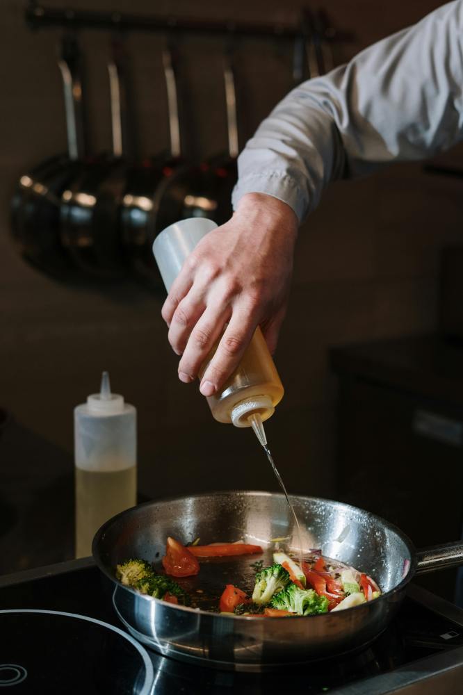 Person pouring oil into frying pan with broccoli and tomato pieces on stove.
