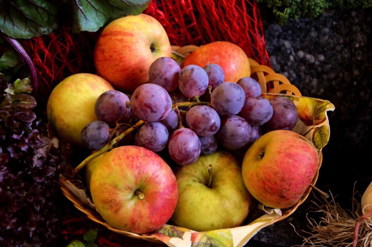 Basket with red apples and purple grapes surrounded by leaves.