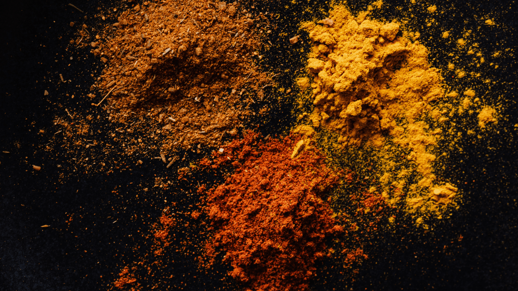 Three piles of spices in brown, yellow, and red on a dark surface.