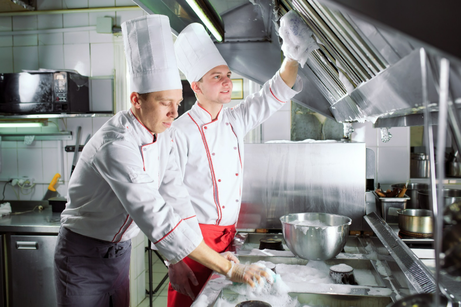 Two chefs cleaning a kitchen hood, wearing white uniforms and hats in a professional kitchen setting.