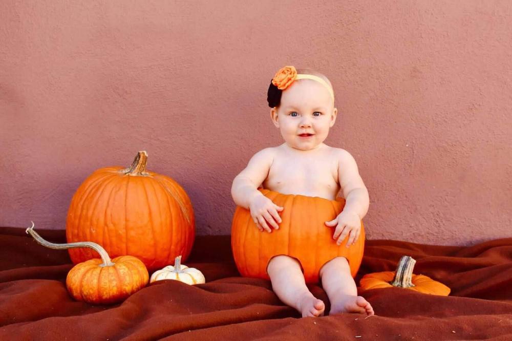 Baby sitting in a pumpkin, surrounded by three pumpkins on a blanket, wearing a flower headband.