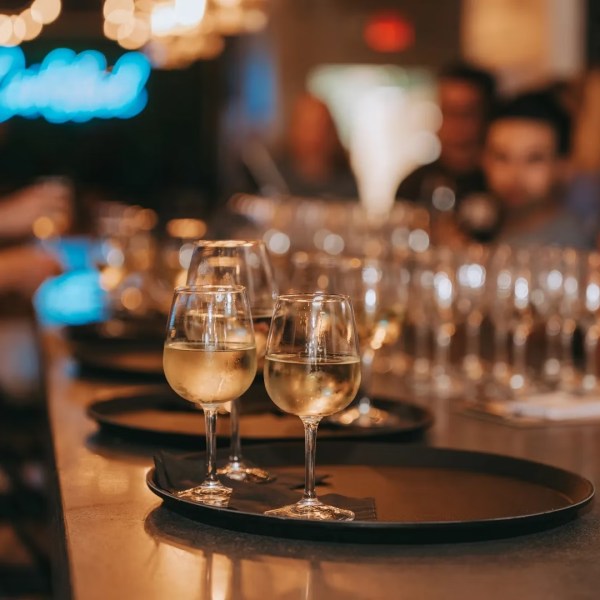 Wine glasses on tray at bar with blurred patrons and blue neon sign in background.