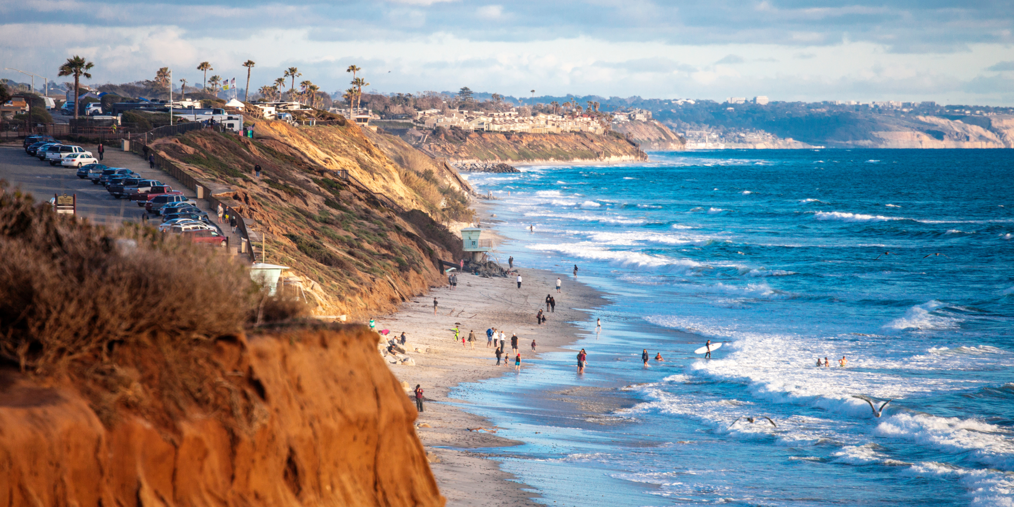 Coastal view with cliffs, beachgoers, and waves at a sandy shore under a cloudy sky.