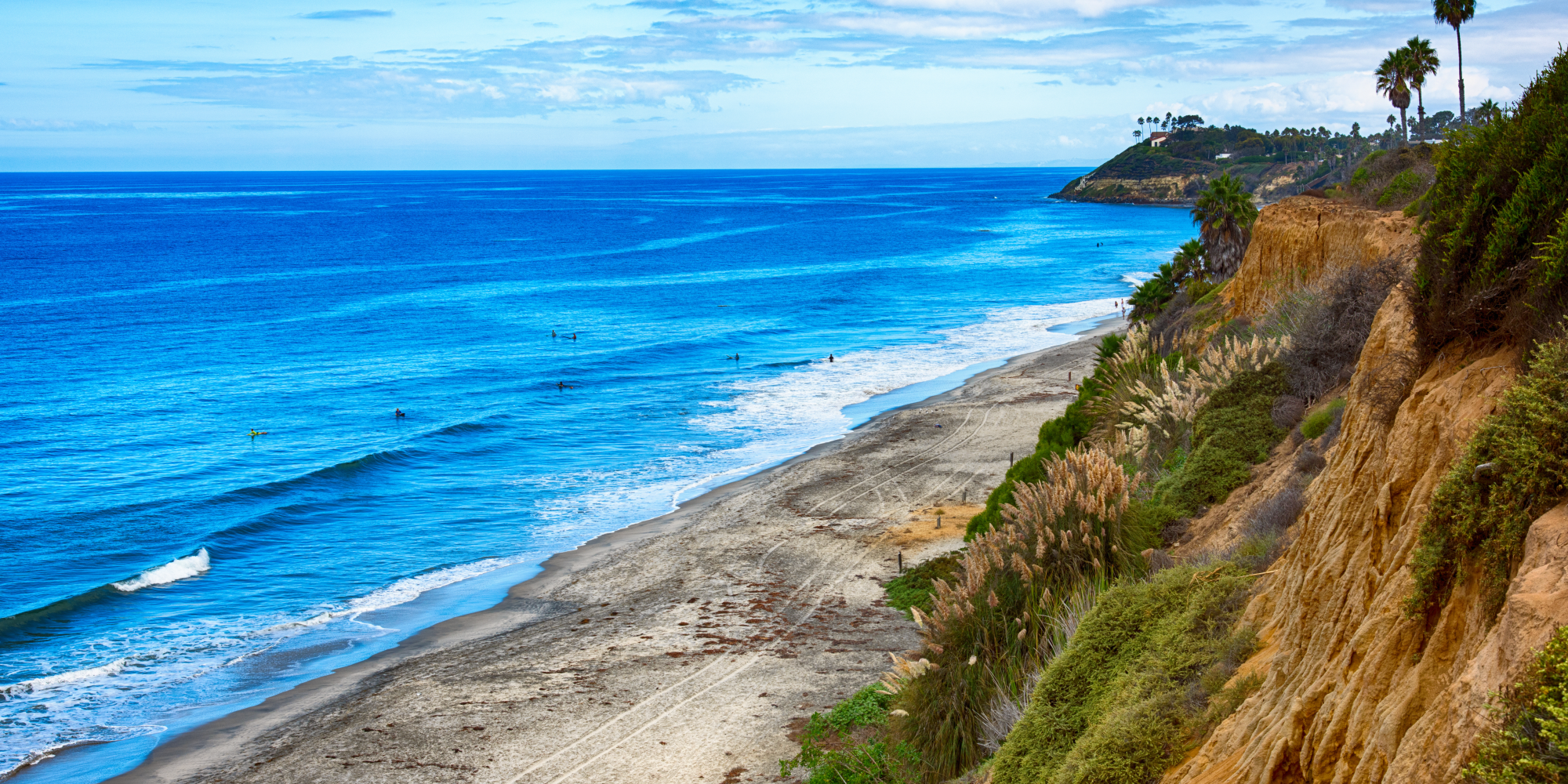 Carlsbad Beaches Coastal view with blue ocean, sandy beach, and rocky cliffs, under a partly cloudy sky.