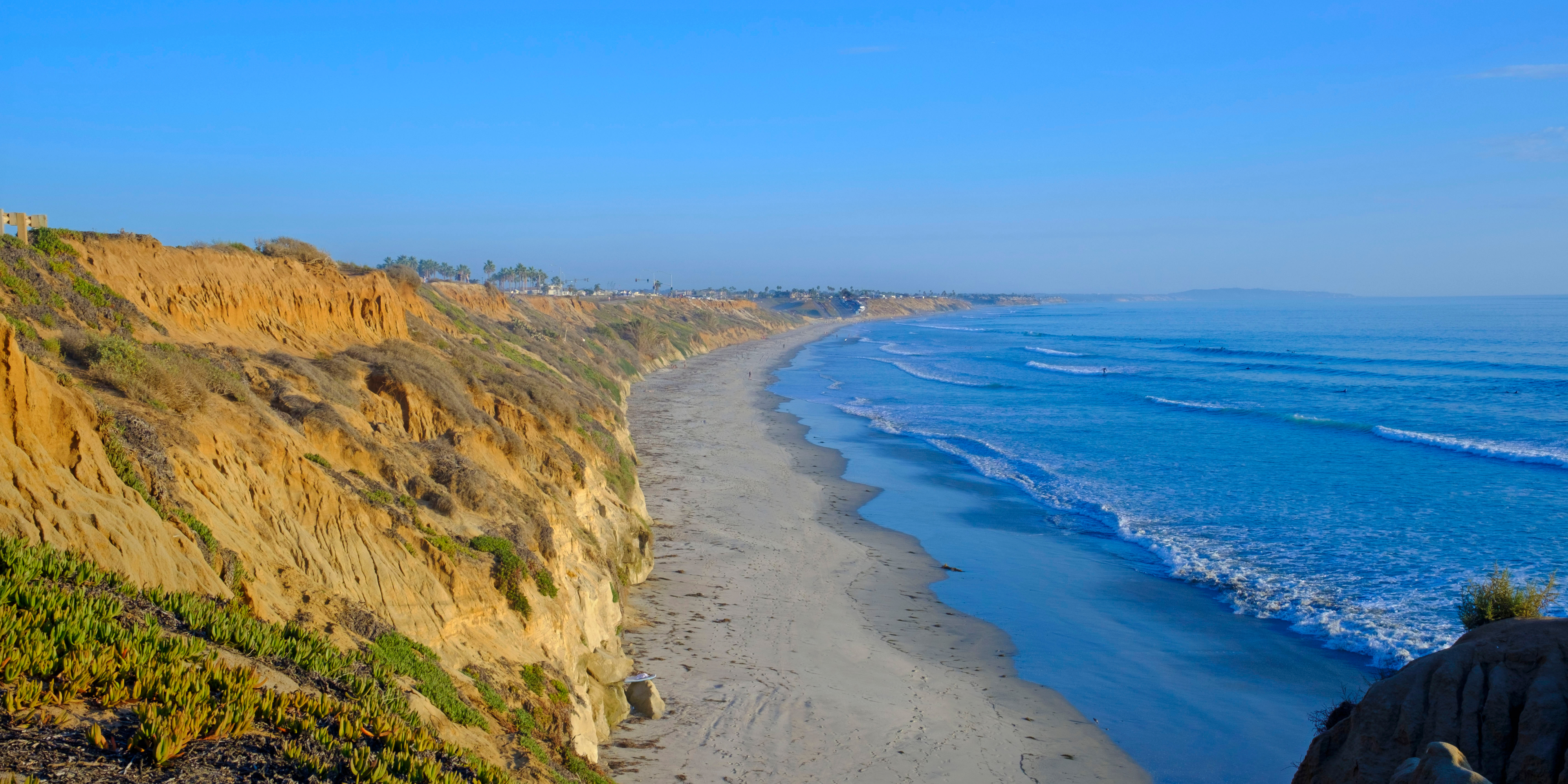 Coastal view with sandy beach, cliffs, and ocean waves under a clear blue sky.