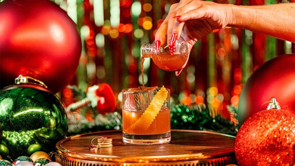 Hand pouring liquid into glass with orange peel, surrounded by Christmas ornaments.