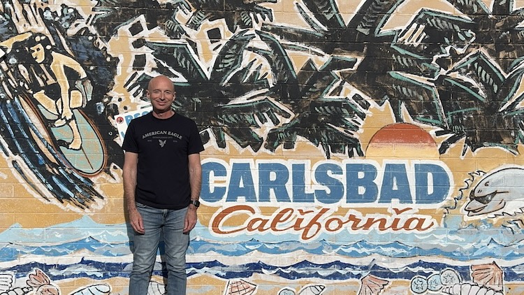 Man standing beside a mural of palm trees and ocean with 'Carlsbad California' text.