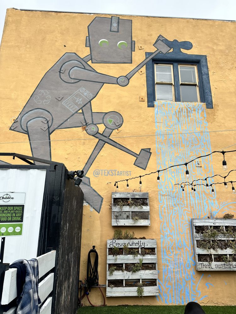 Mural of a robot painting a wall with windows, plants, and text about sustainability on a yellow building.