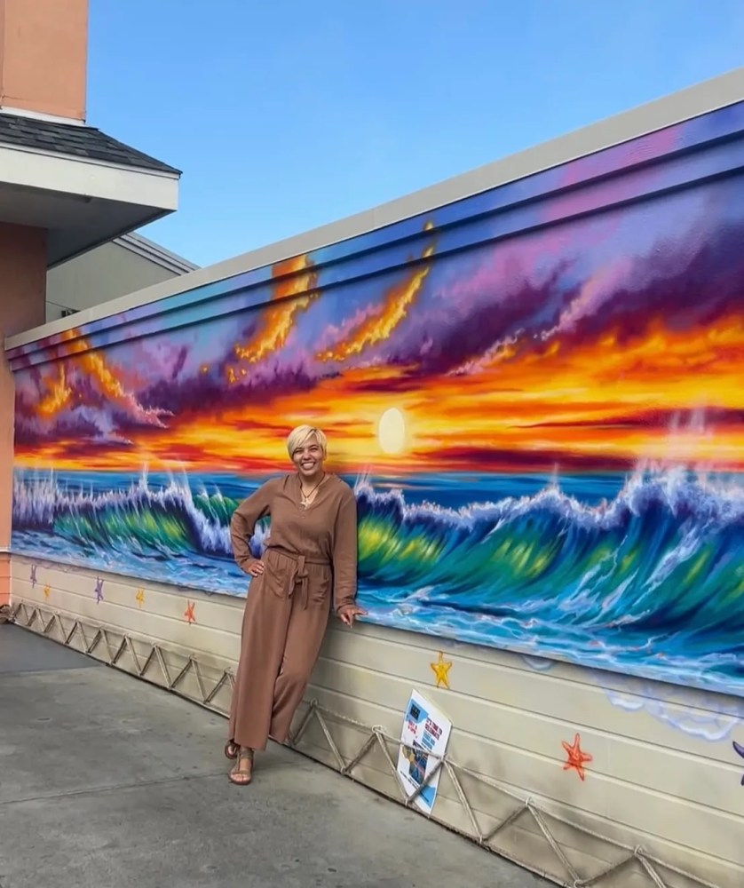 Person in brown outfit poses by colorful ocean wave mural under a blue sky.