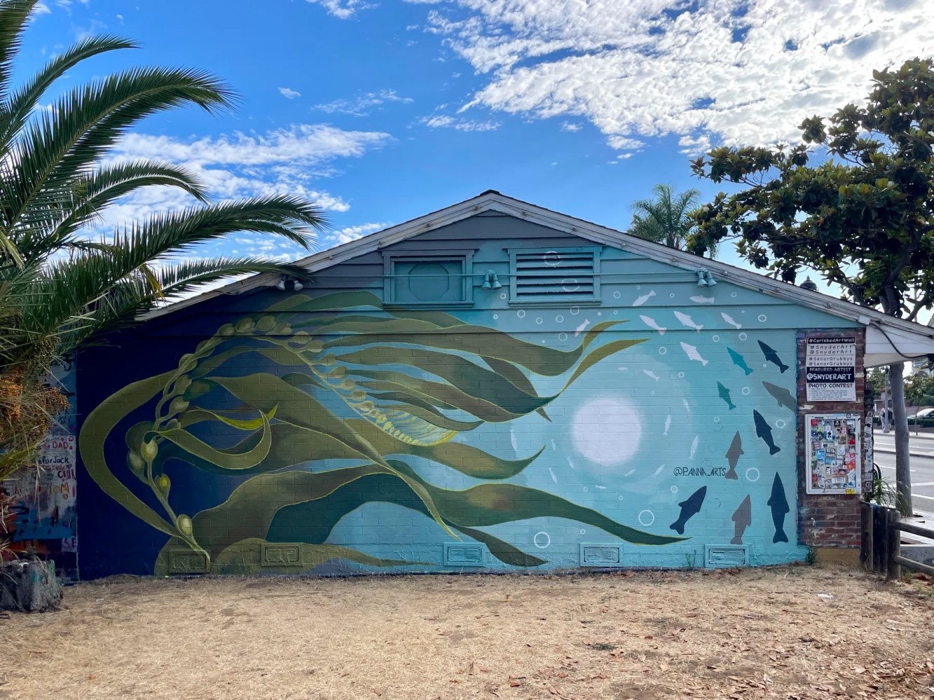 Mural on building wall depicts underwater scene with kelp and fish silhouettes under a blue sky.