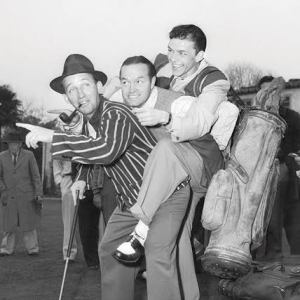 Three men on a golf course, one in a hat holding a club, another being playfully carried.
