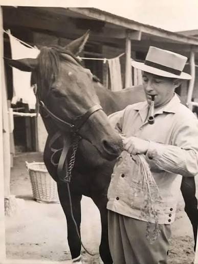 Person in a hat feeding a horse with a pipe in mouth, outdoor stable setting.