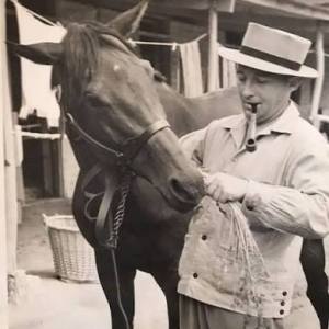 Person in a hat feeding a horse with a pipe in mouth, outdoor stable setting.