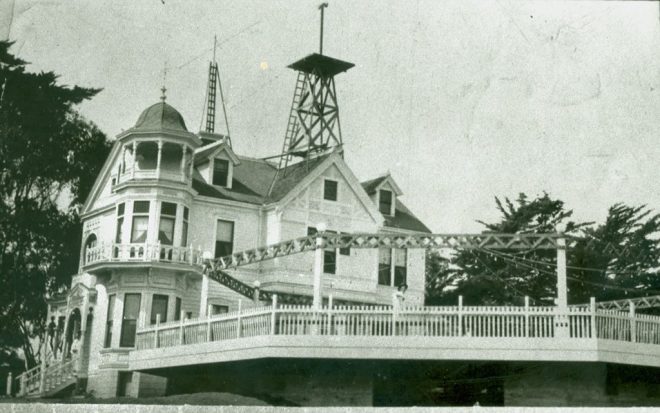 Black and white photo of large Victorian house with a tower and surrounding trees.