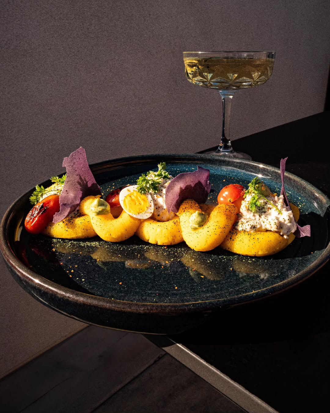 Elegant plated dish with deviled eggs, tomatoes, and herbs, plus a glass of champagne on a dark surface.
