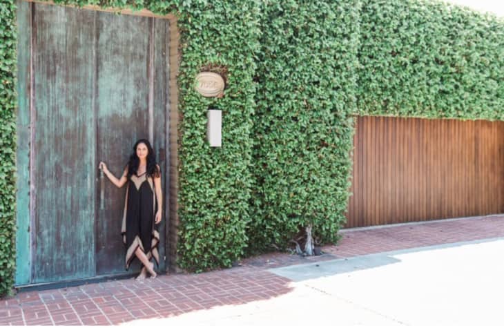Person in black dress stands by large green door, ivy-covered wall, and wooden gate.