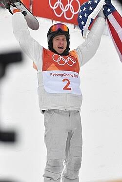 Snowboarder in Olympic gear holding a snowboard and American flag.
