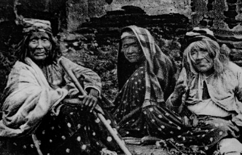 Three women in traditional attire sitting outdoors, holding walking sticks.