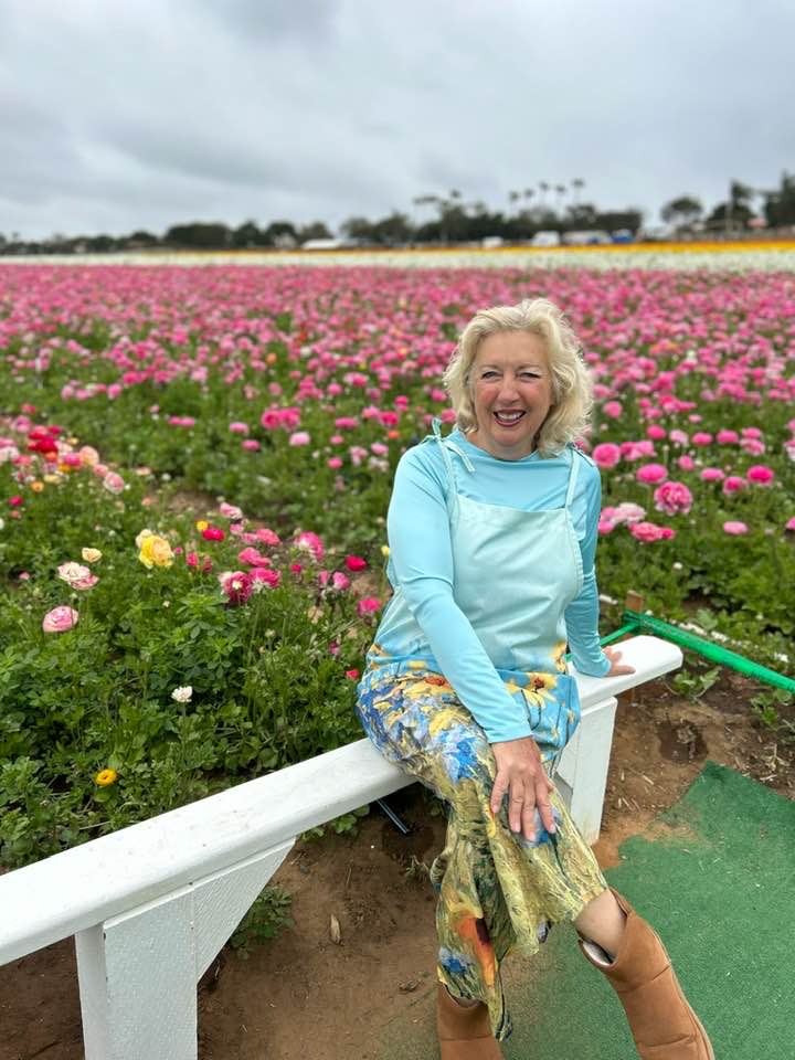 Smiling person in a floral field, wearing a light sweater and floral skirt.