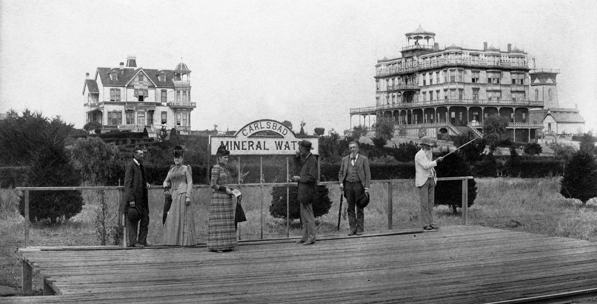 Victorian people standing on a boardwalk near a mineral water sign with old buildings in the background.