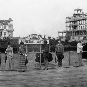 Vintage photo of people by 'Mineral Water' sign with large buildings in the background.