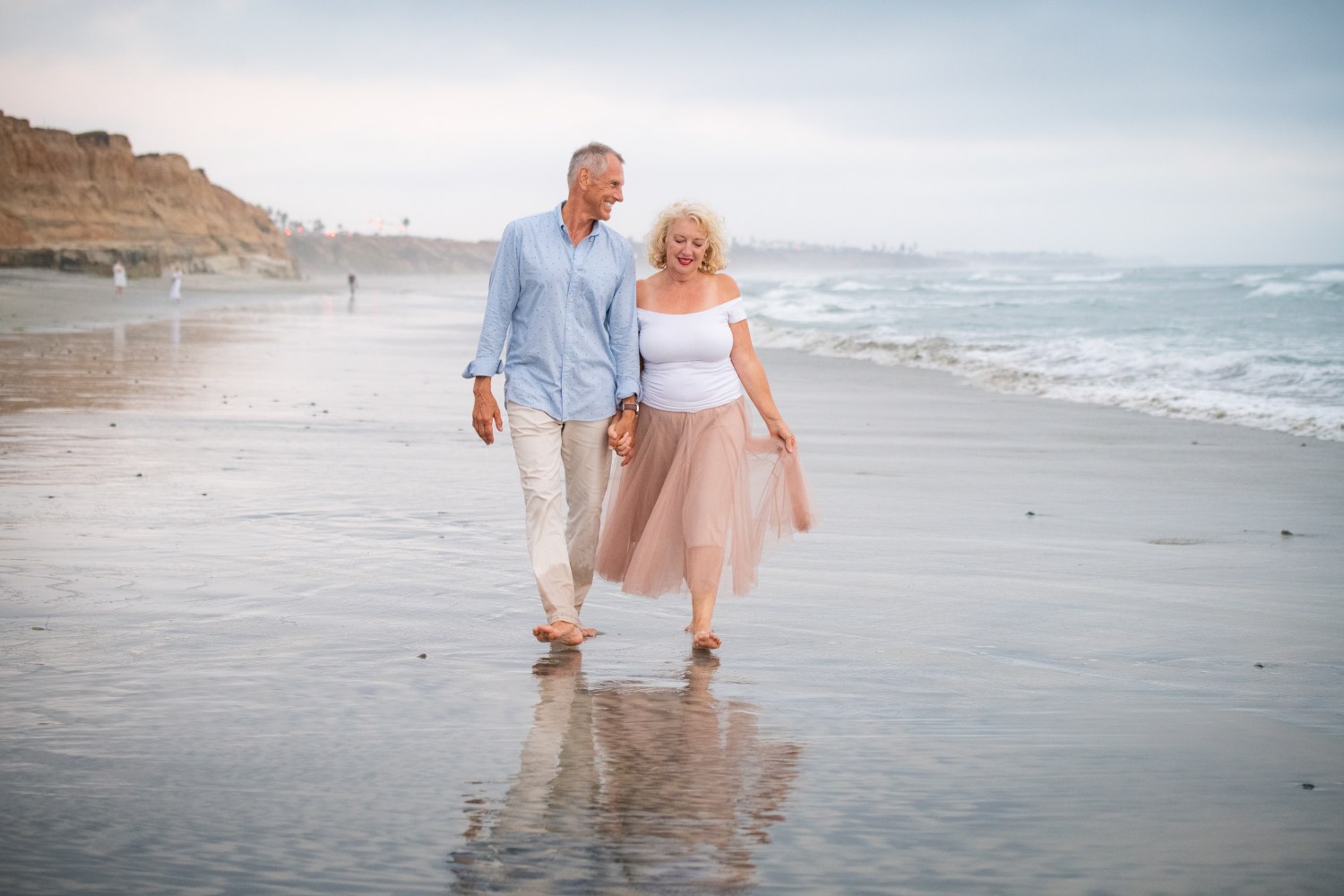 Older couple holding hands walking along a sandy beach.