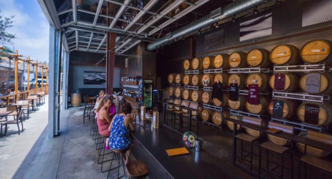 Open garage-style brewery with patrons sitting at a bar next to displayed barrels.