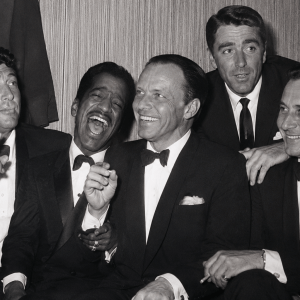 Five men in tuxedos laughing and smoking, sitting close together.