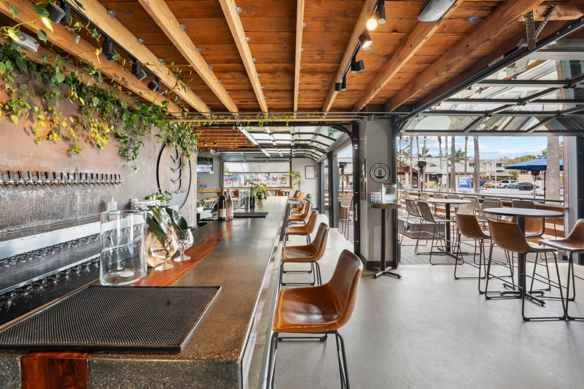 Modern bar interior with high chairs, wooden ceiling, and open glass doors to patio seating.