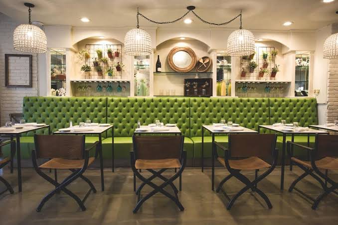 Chic restaurant with green tufted seating, brown chairs, and hanging lights.