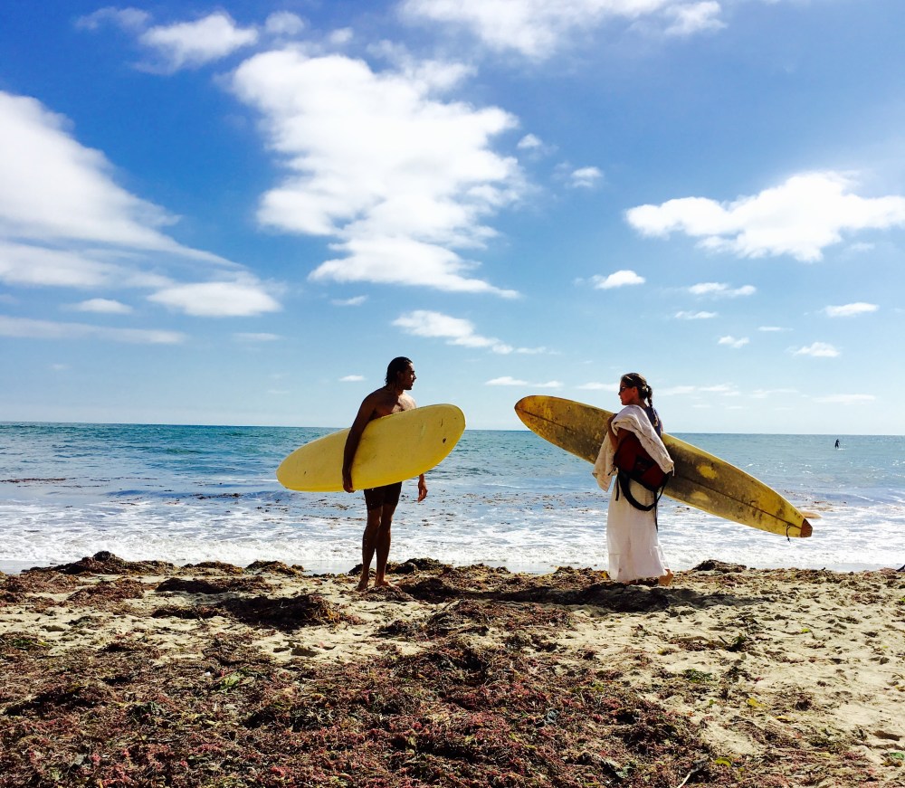 Two surfers with yellow boards stand on a sandy beach under a blue sky.