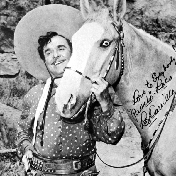 Man in cowboy attire smiling, standing beside a horse with a rocky background.