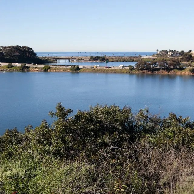 Scenic view of a lake with greenery in foreground, hills, and blue sky.