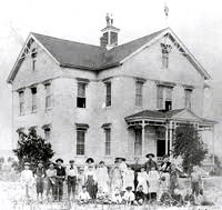 Vintage photo of people in front of a large brick house with a porch and dormer windows.